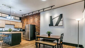 A modern kitchen with a brick wall, wooden flooring, and a dining area with a table and chairs. - 2BR-1BA, Sleeps 4 | Downtown Ames (Ames)