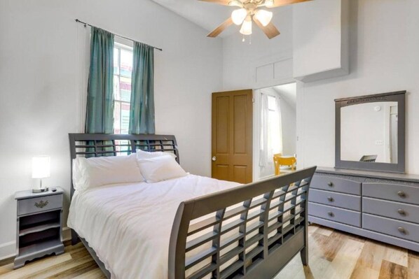 Un dormitorio con cama, mesita de noche, cómoda, espejo, ventilador de techo y una puerta.