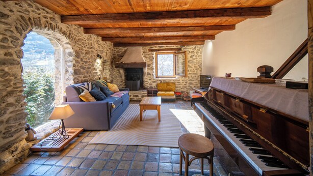 A rustic stone-walled room with a wooden ceiling, a sofa with cushions, a piano, a fireplace, and a view through an arched window. - Mountain house near Méribel (Brides-les-Bains)