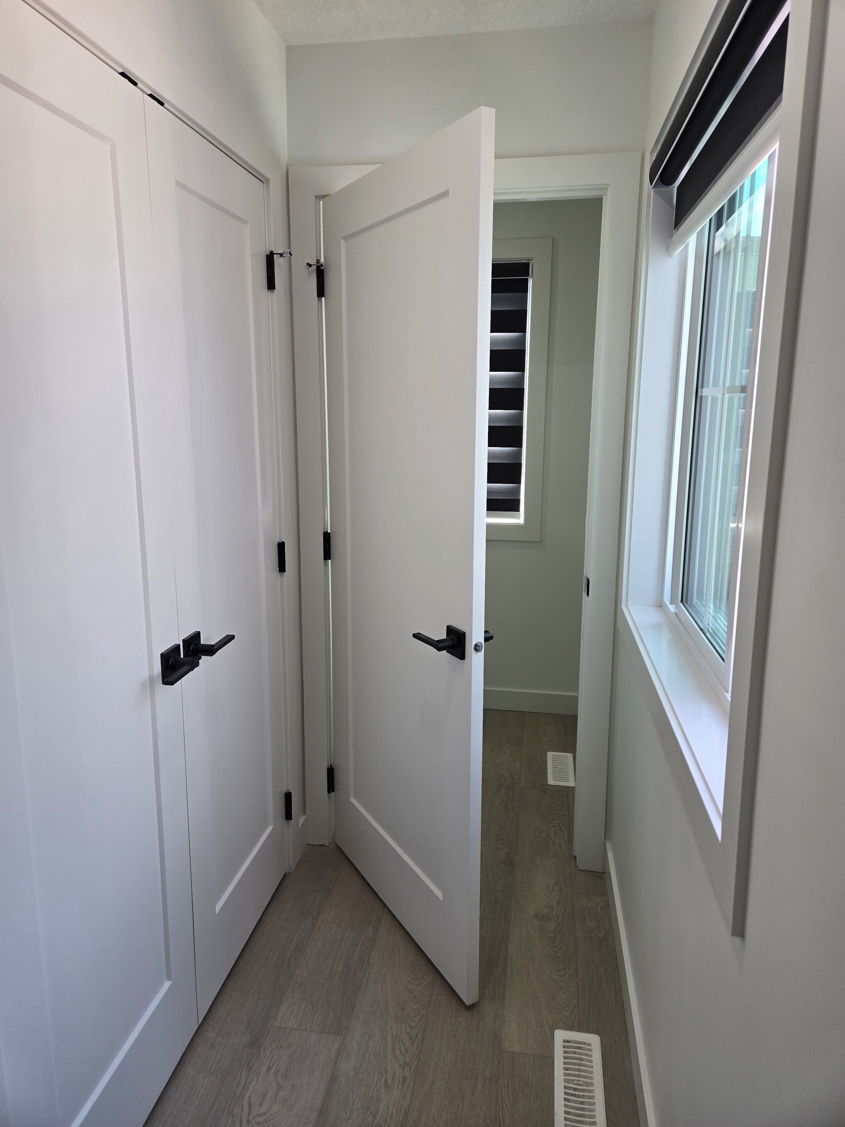 A hallway with white doors, a window with blinds, and a vent on the wall.