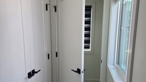 Un pasillo con puertas blancas, un ventanal con persianas y una rejilla en la pared.