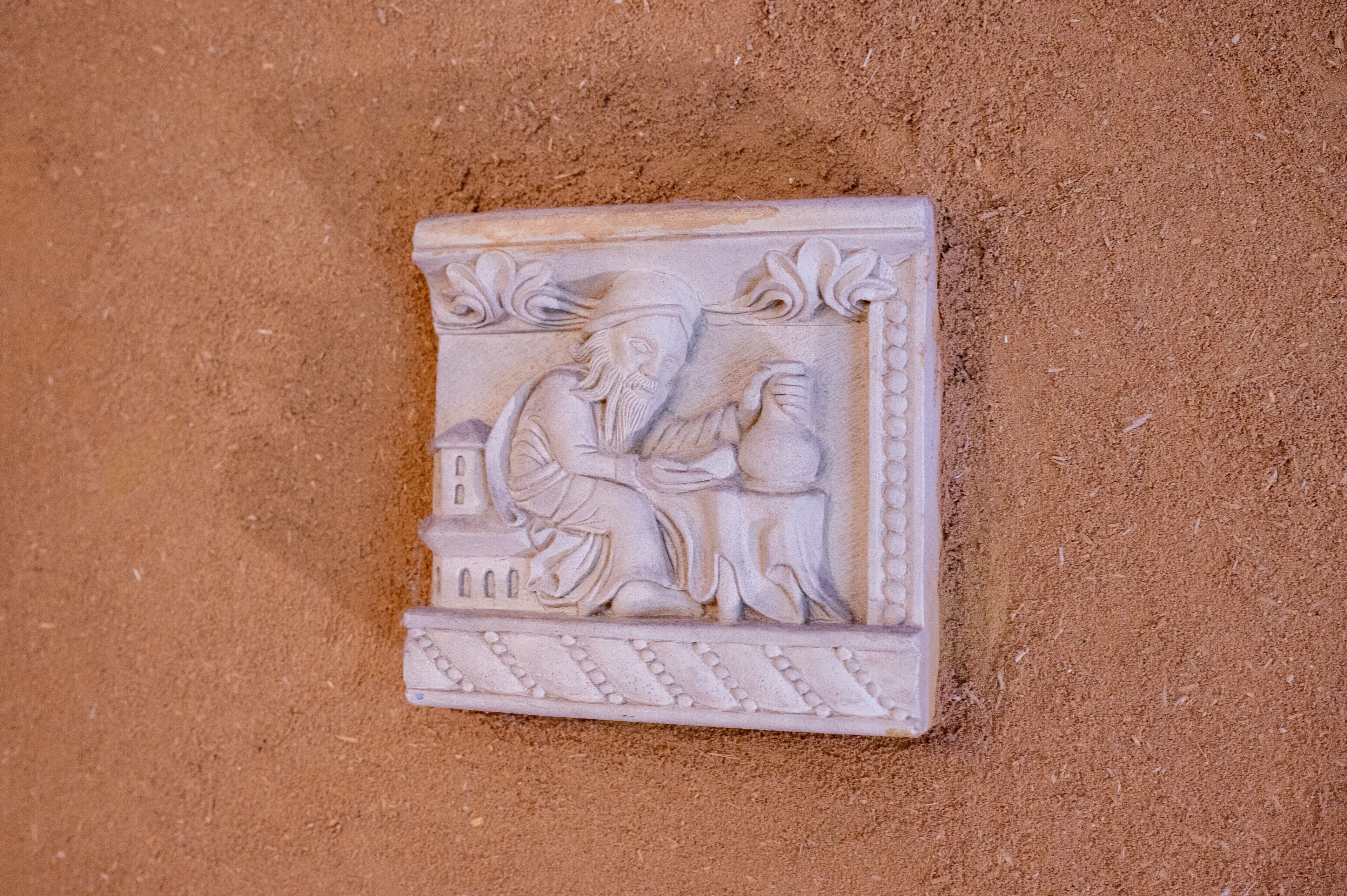 Een stenen reliëfbeeldhouwwerk met een zittend figuur en een ander figuur in profiel, voor een zandachtige achtergrond.