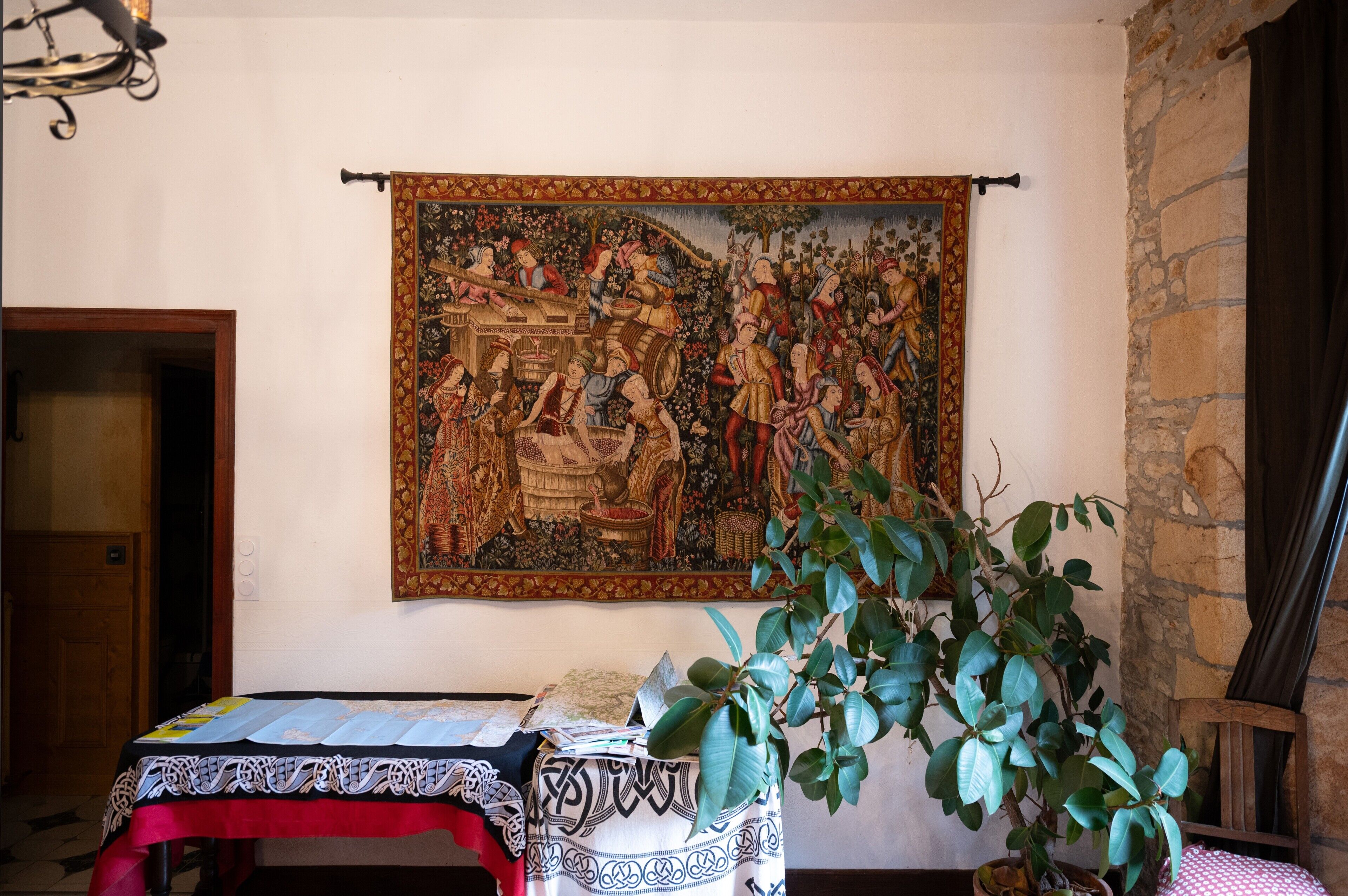 A room with a tapestry, a table with a patterned tablecloth, a potted plant, and a stone wall.