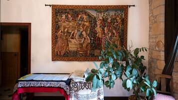 A room with a tapestry, a table with a patterned tablecloth, a potted plant, and a stone wall.