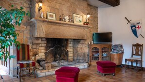 A stone fireplace with a television above it, two red ottomans, a wooden chair, and a shield on the wall. - Bed and Breakfast 'Perceval' with Shared Terrace, Shared Garden and Wi-Fi (Pluvigner)