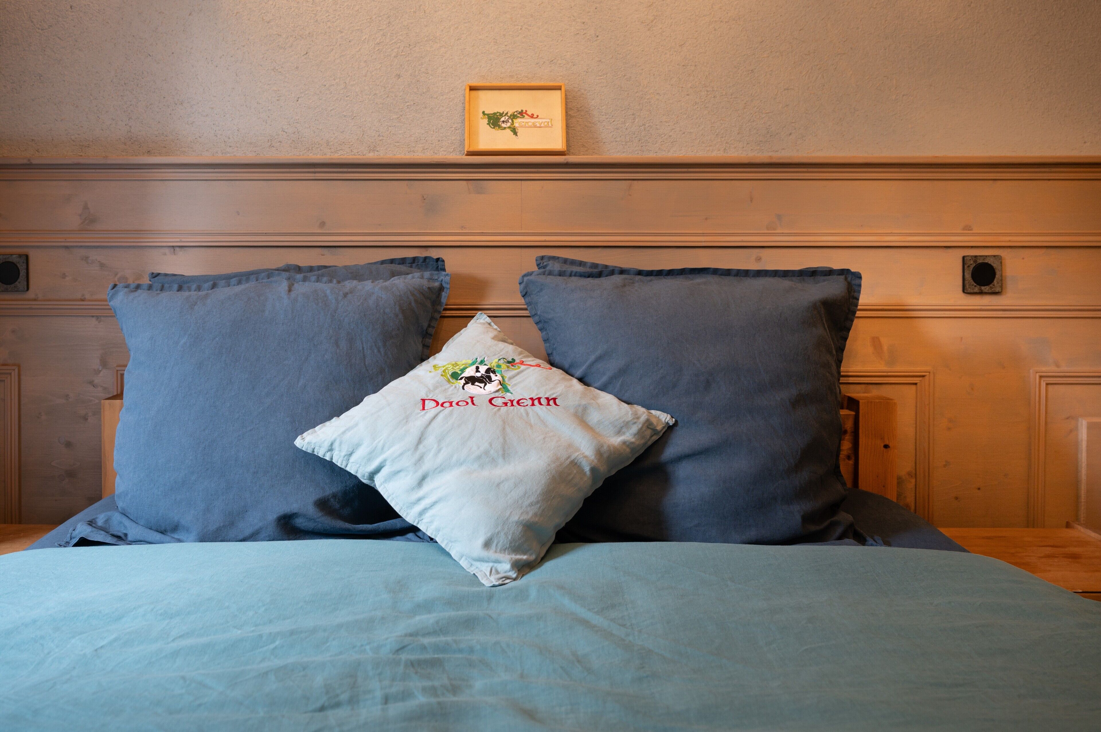 Een bed met een blauwe spreidek en twee kussens, waarvan één een naam heeft.