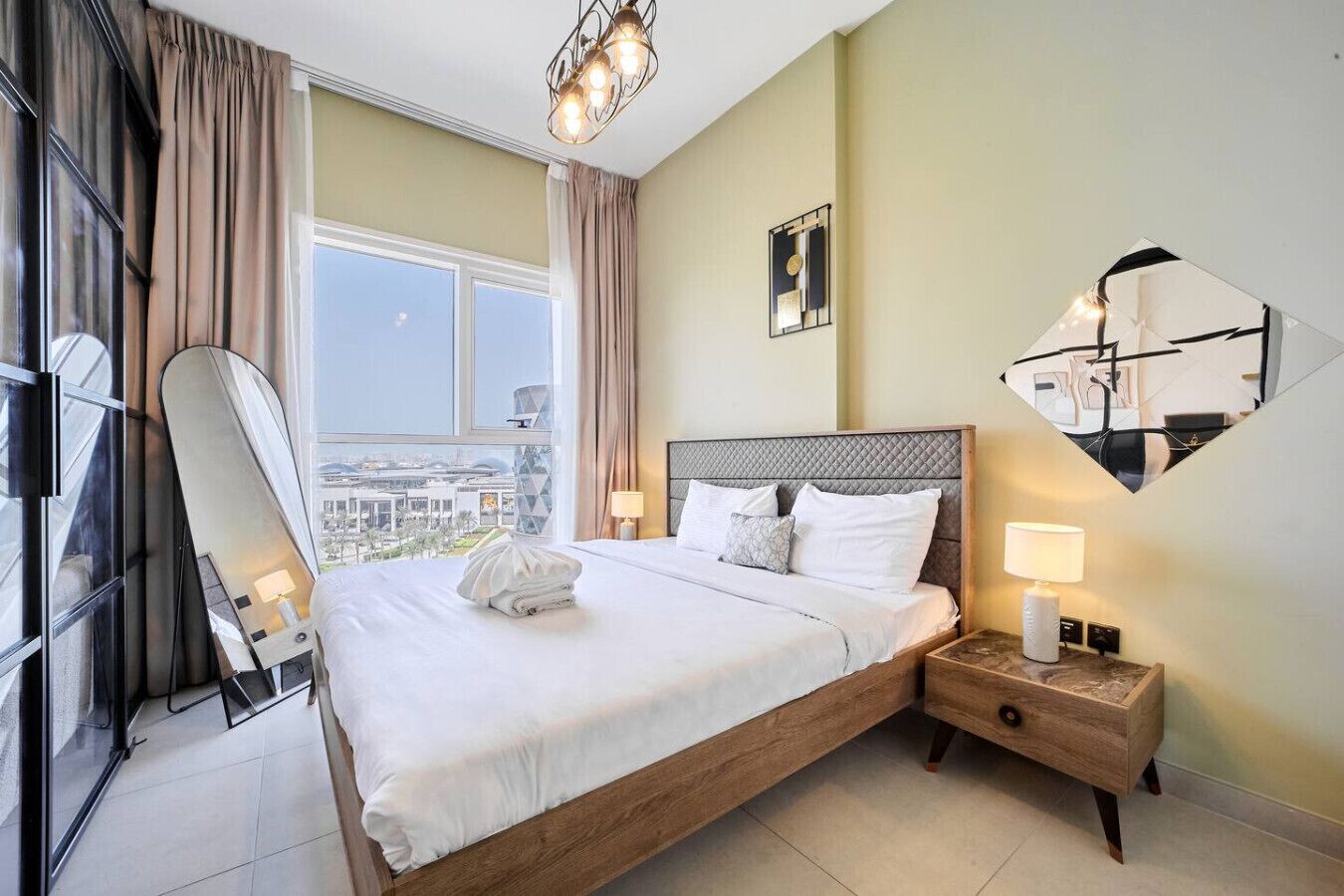 Une chambre d’hôtel avec un grand lit, une table de chevet avec une lampe, un miroir et une vue sur la ville grâce à de grandes fenêtres.