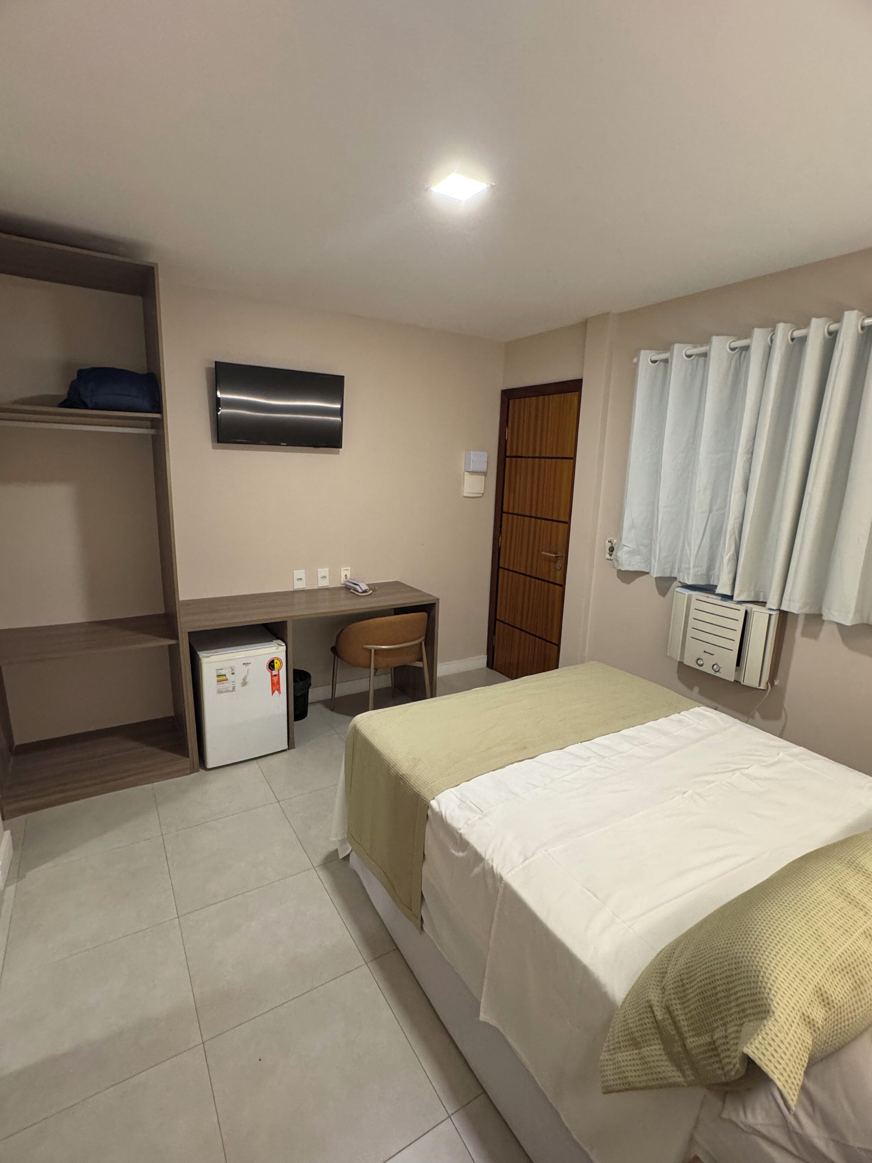 Un dormitorio con cama, escritorio, silla, refrigerador pequeño, televisor y baño con cortina de ducha.
