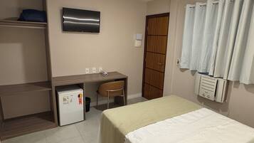 Un dormitorio con cama, escritorio, silla, refrigerador pequeño, televisor y baño con cortina de ducha.