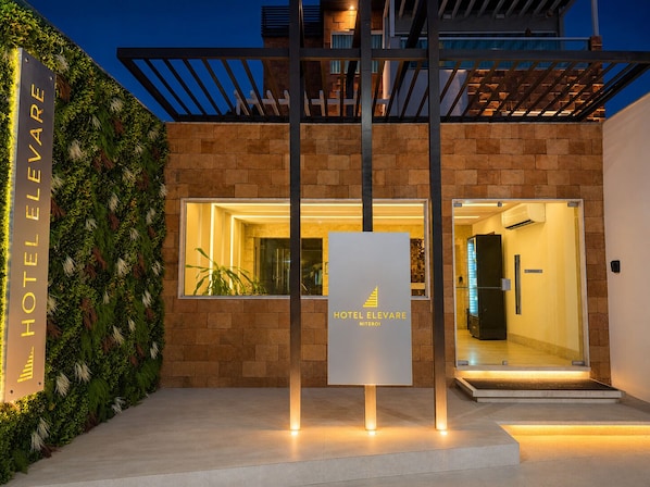 Une entrée d’hôtel moderne, avec une enseigne indiquant « HÔTEL ELEVARE NITEROI », une façade en briques et une porte en verre donnant sur l’intérieur.