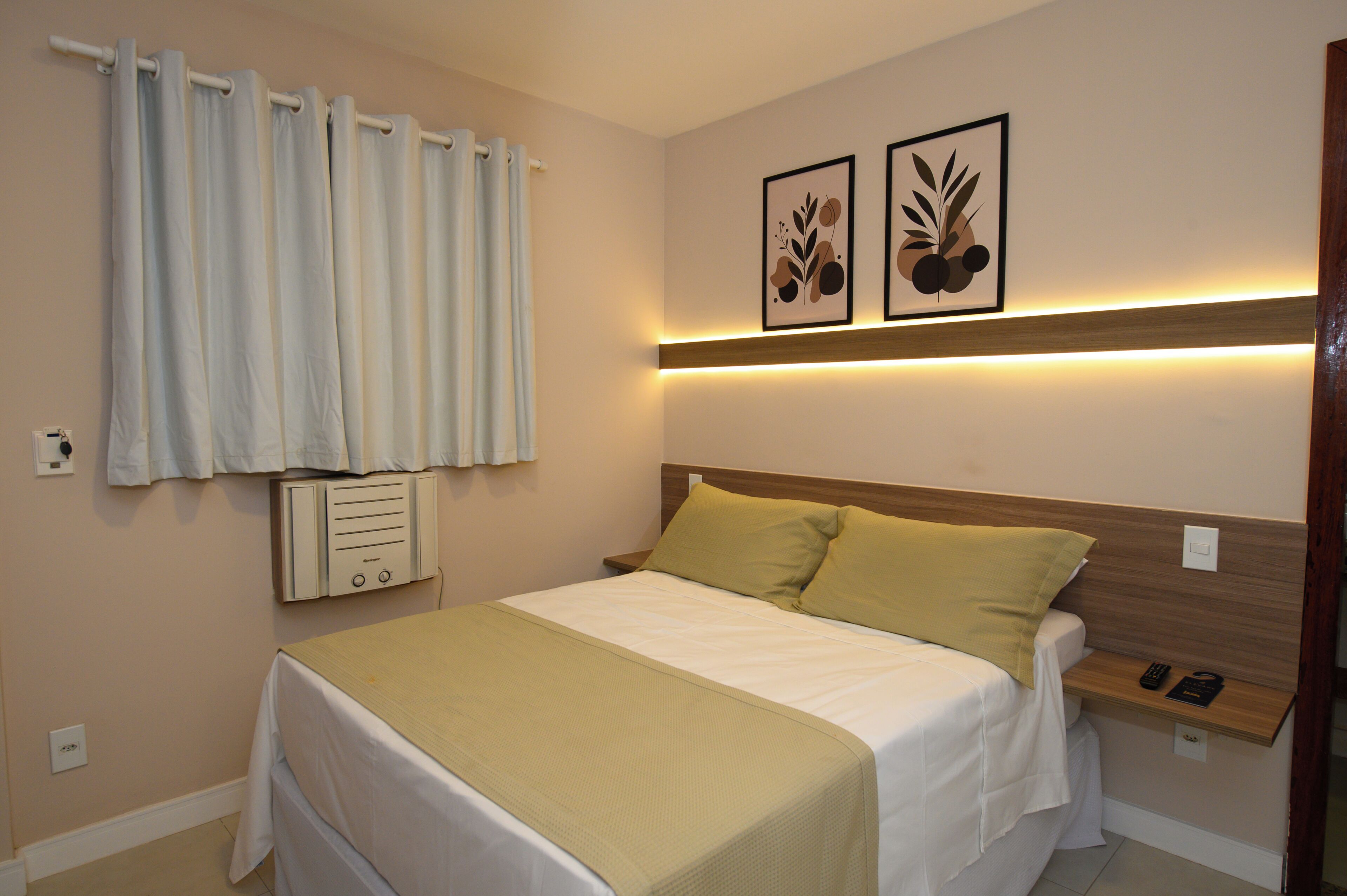 A neatly made bed with a wooden headboard, a bedside table with a remote control, and a wall-mounted air conditioning unit.