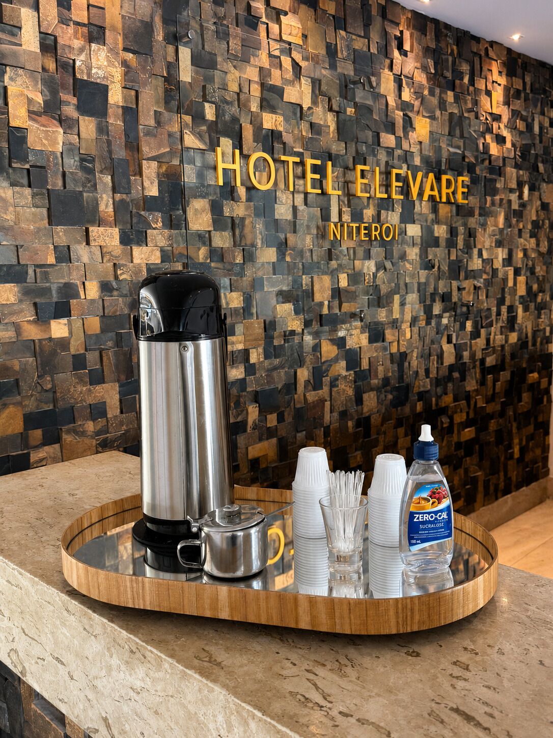 Zona de recepción de hotel con pared de piedra, una máquina de café y una bandeja con tazas y una botella de agua.