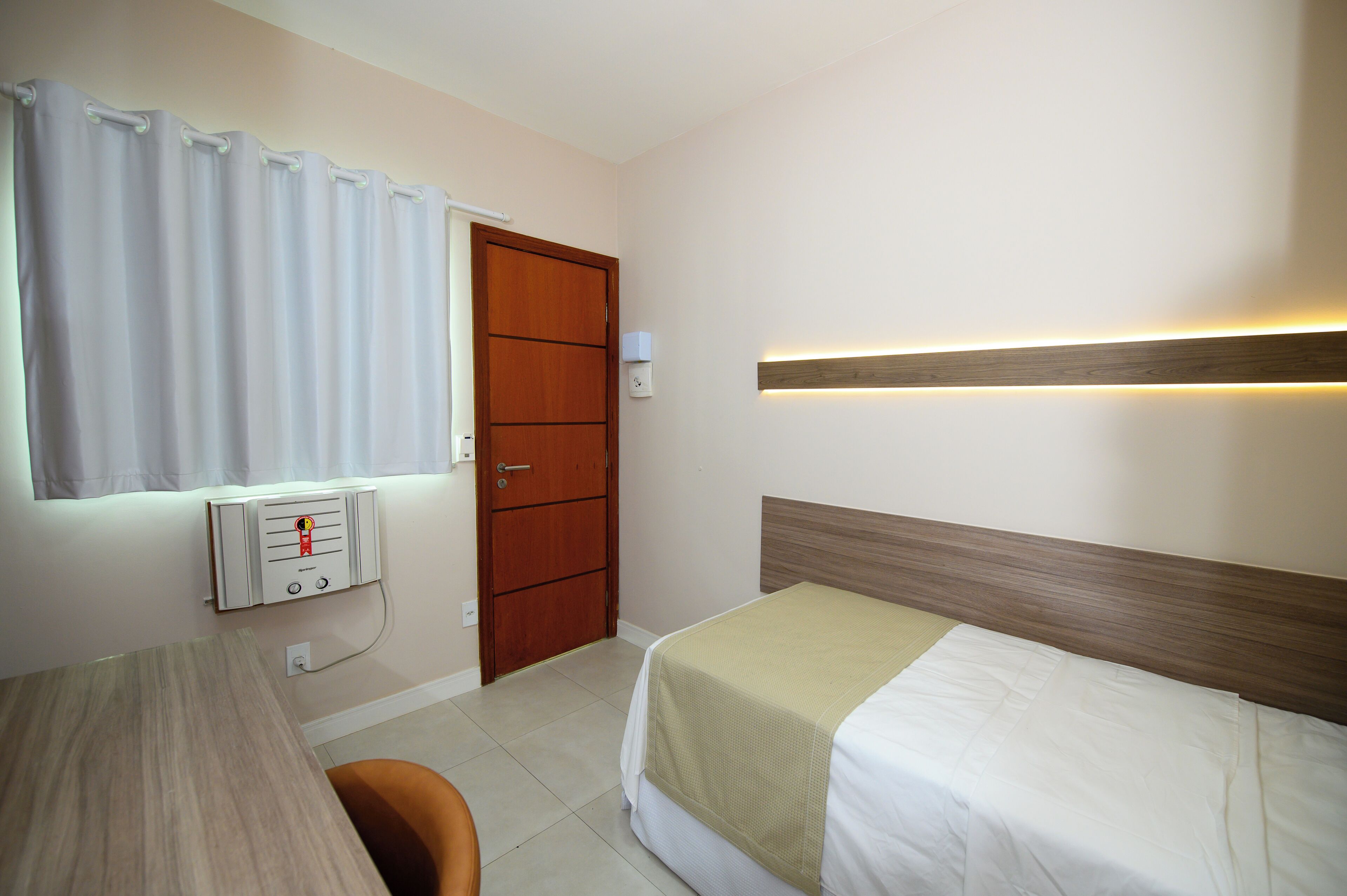 A hotel room with a bed, a desk, a chair, a window with curtains, a door, and a wall-mounted light fixture.