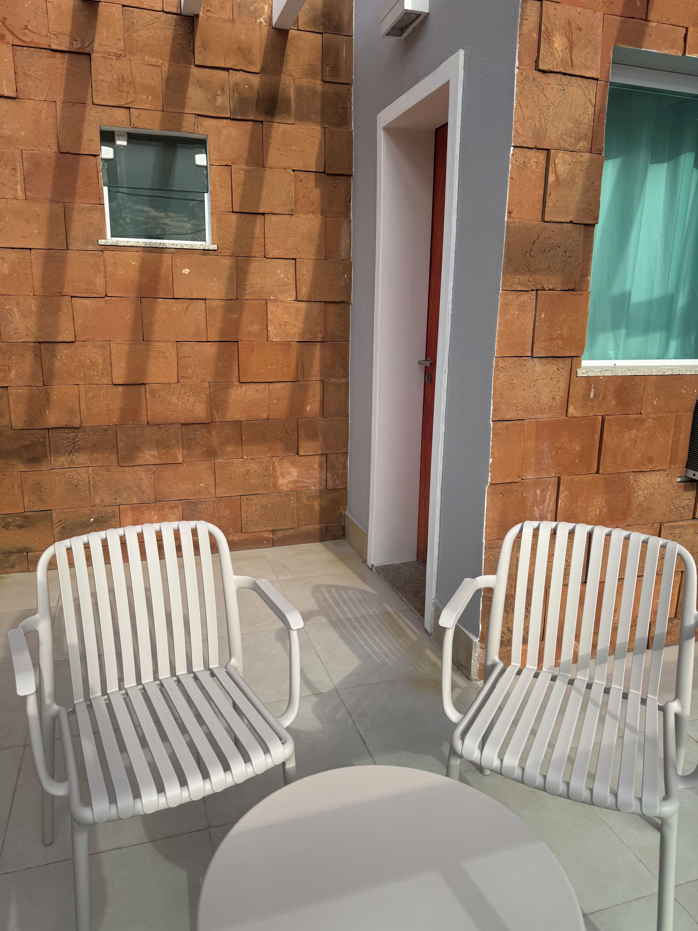 A patio area with two white metal chairs and a small round table, a brick wall, a door, and a window with a curtain.