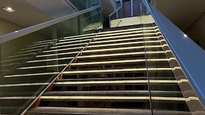 Una escalera moderna con barandillas de vidrio y escalones iluminados, que asciende en un edificio con techo de cristal.