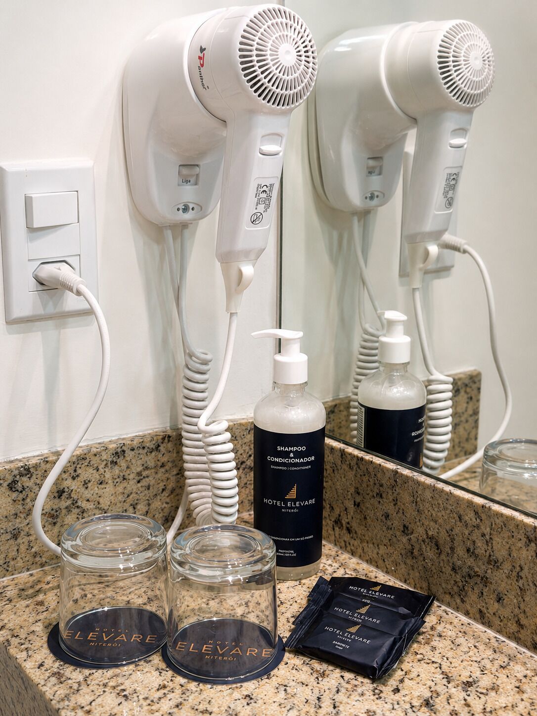 A hotel bathroom with two hair dryers, a bottle of shampoo, and two glass jars on the counter.