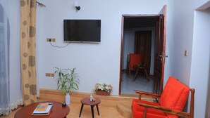 Ett vardagsrum med en platt-TV som är monterad på väggen, en trästol med en orange kudde, ett litet runt bord med en bok och en flaska, och en krukväxt.