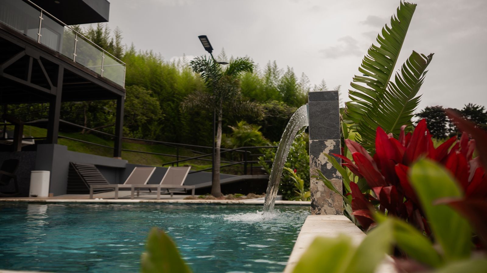 Ein Schwimmbecken mit Wasserfall, Liegestühlen und einem modernen Gebäudebau im Hintergrund.