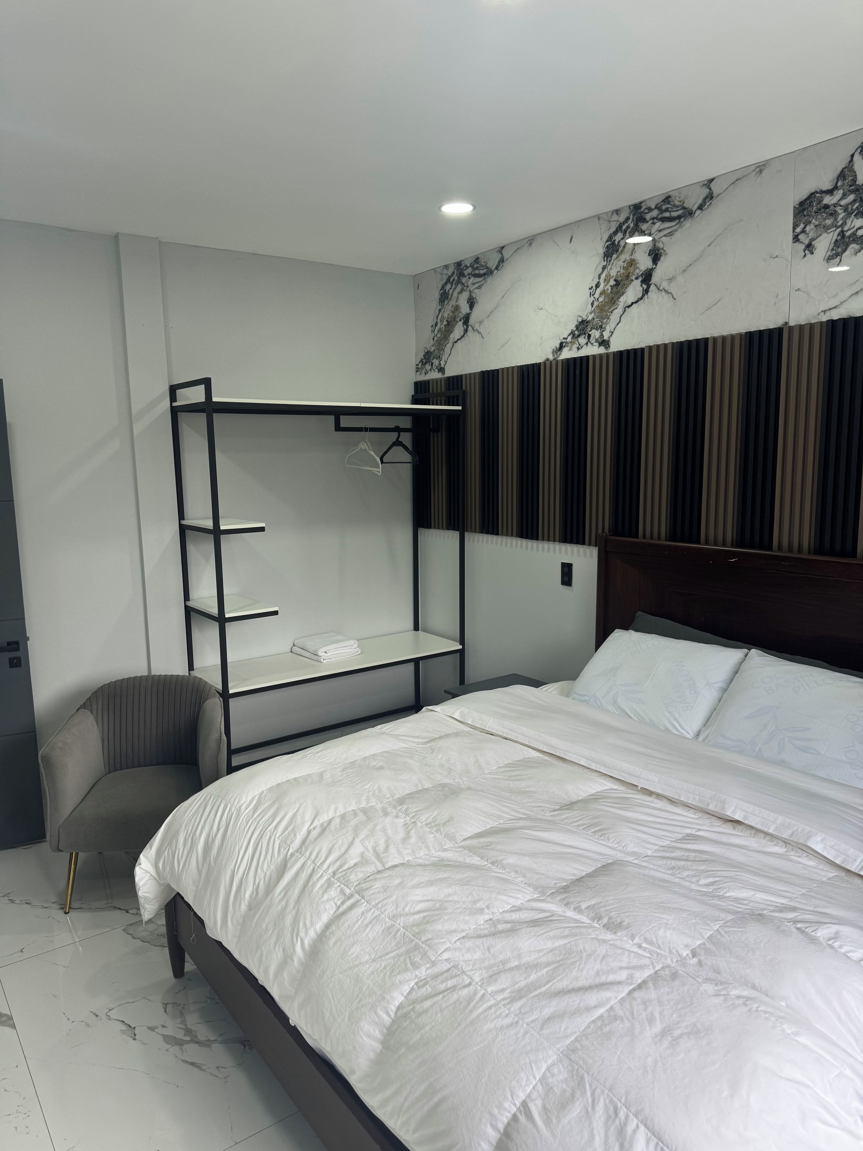 A modern bedroom with a large bed, a built-in wardrobe, a chair, and a marble-patterned wall.