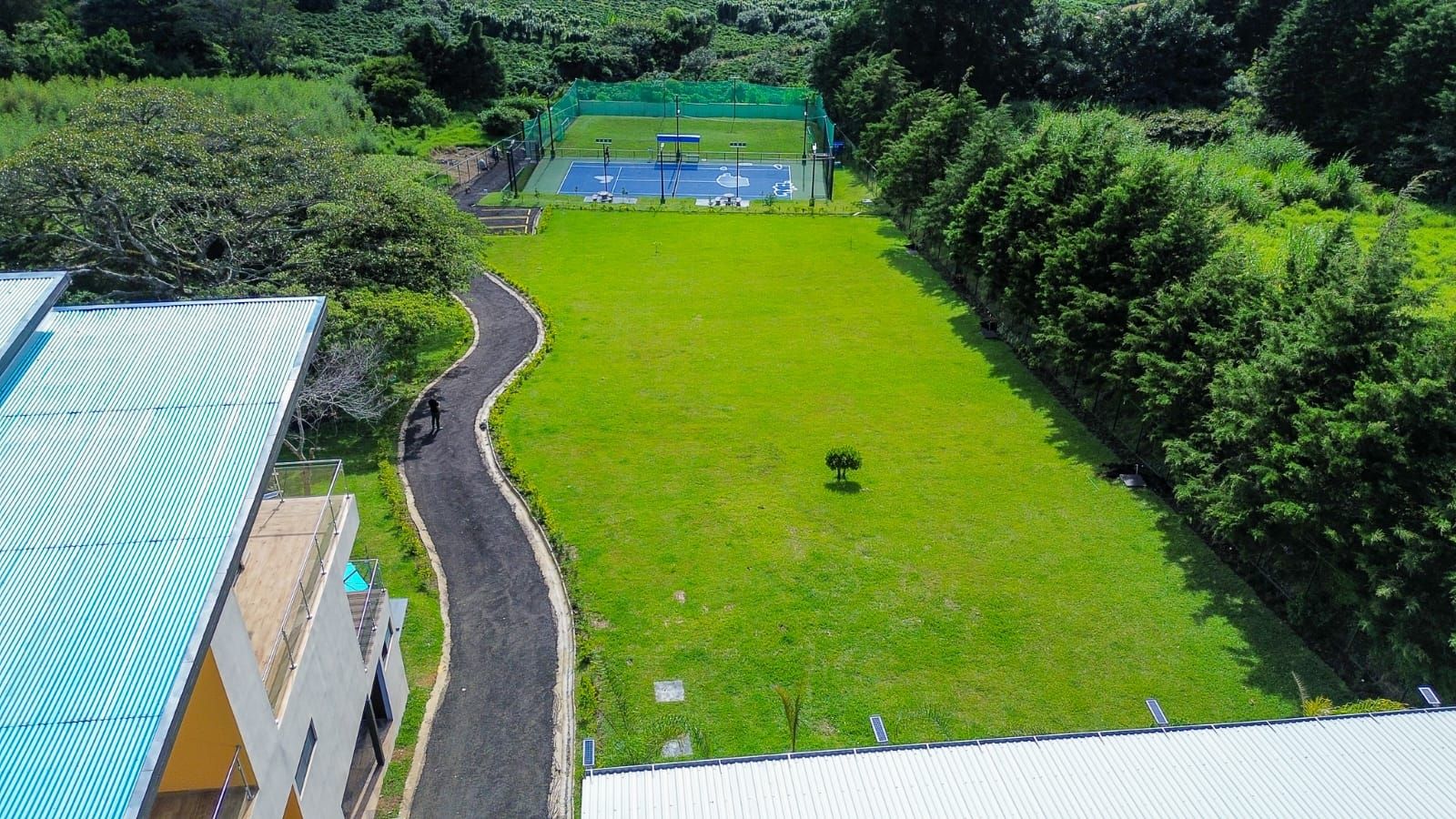 An aerial view of a sports court surrounded by lush greenery, with a road curving past a building with a green roof.