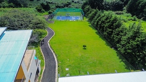 An aerial view of a sports court surrounded by lush greenery, with a road curving past a building with a green roof. - Luxury Home Gym Jacuzzi Pool Suana Soccer Field and Tennis & Basketball Courts (Heredia)