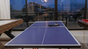 Une table de ping-pong se trouve dans une pièce dotée de grandes fenêtres, avec une table en bois à gauche et une table rouge à droite.