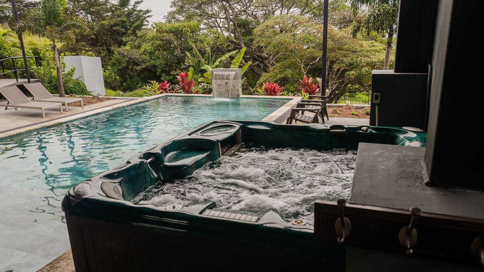 Un bain à remous agrémenté d’une cascade, entouré d’une piscine et de chaises longues, avec une végétation luxuriante en arrière-plan.