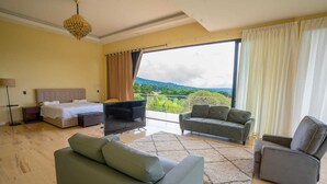 Una sala de estar amplia con una cama grande, un sofá, una silla y un televisor. Cuenta con un balcón con vistas a las montañas.