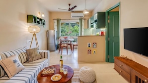 A living room with a sofa, coffee table, and a TV. There is a dining area with a table and chairs, and a kitchen with cabinets and a TV. - 1BR in North Goa Pool Gym Clubhouse Access (Mapusa)