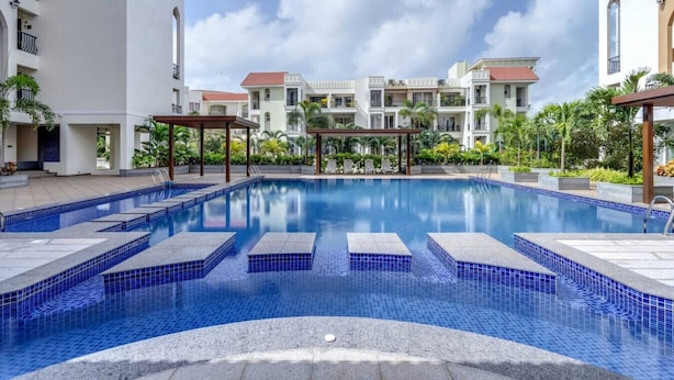 A swimming pool with tiled steps, surrounded by multi-story buildings with balconies and a clear sky. - 1BR in North Goa Pool Gym Clubhouse Access (Mapusa)