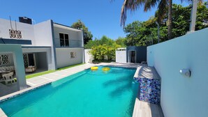Un área de piscina moderna con piscina azul cristalina, dos flotadores amarillos y una pared exterior blanca con detalle de azulejos.