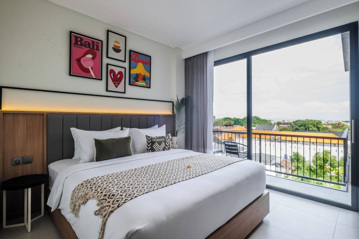 A modern hotel room with a large bed, a view of the city, and Bali-themed artwork on the wall.