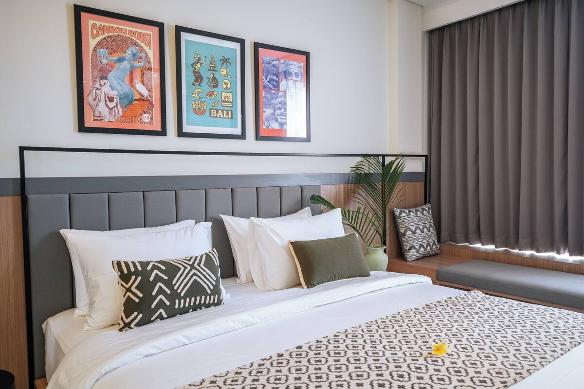 A hotel room with a bed, a headboard, a window with curtains, and three framed pictures on the wall.