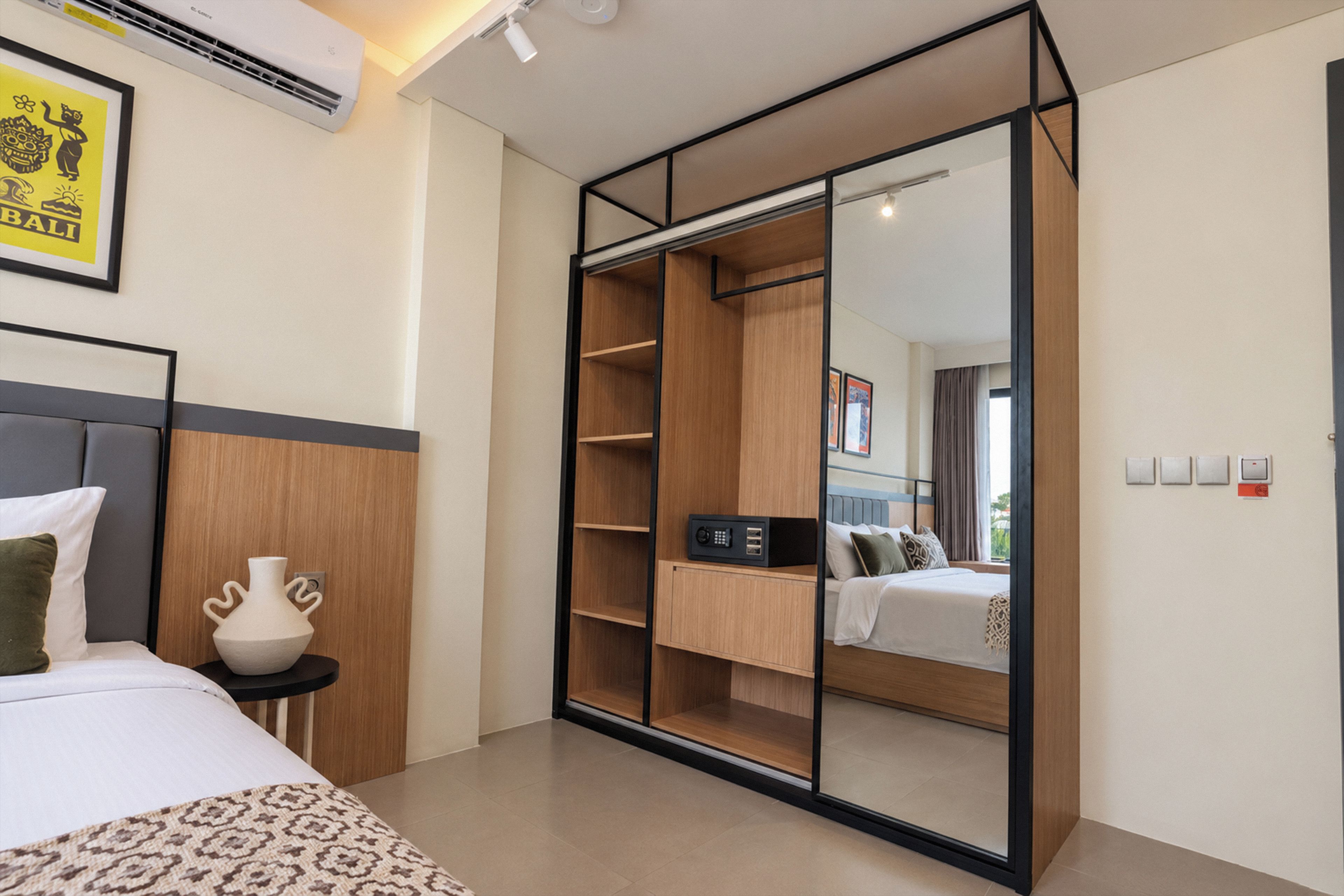 Habitación de hotel con cama, mesita de noche con un florero, armario con espejo y vista a otra habitación.