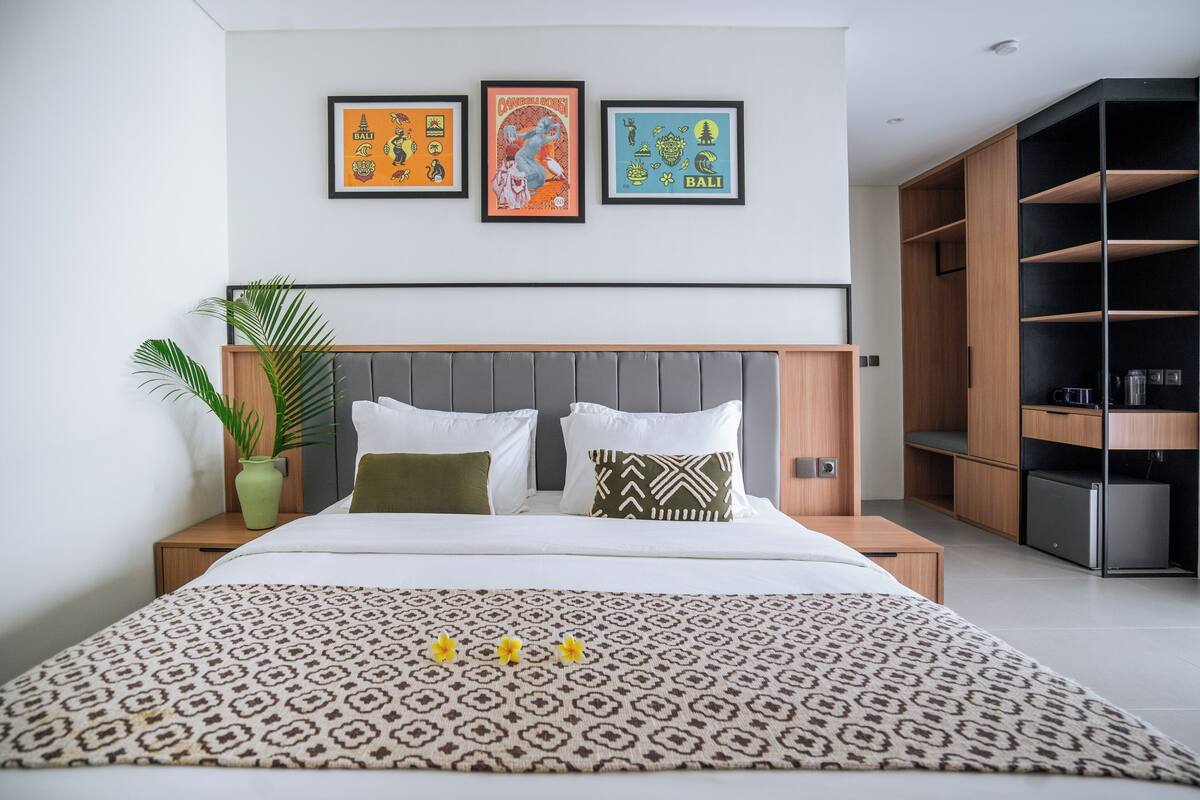 A modern hotel room with a large bed, a nightstand with a plant, a wardrobe, and three framed pictures on the wall.