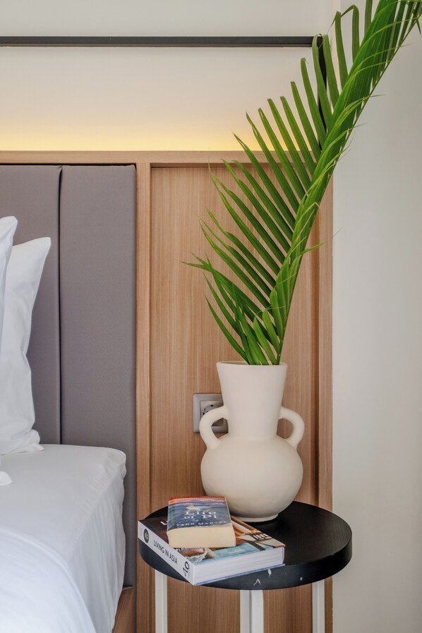 Uma cama bem arrumada com lençóis brancos, uma mesa de cabeceira com um vaso branco e uma planta em vaso, e uma pilha de livros.