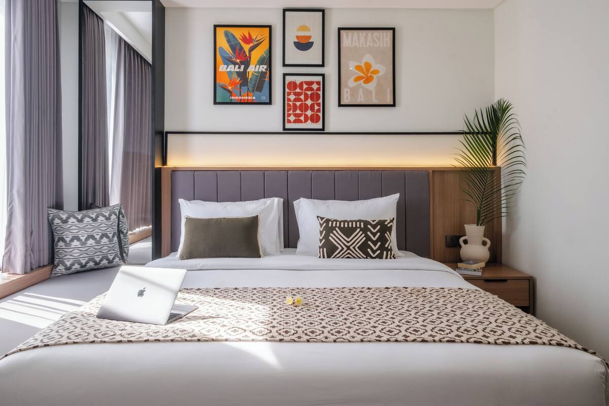 A modern hotel room with a bed, a nightstand, a laptop, and a large window with curtains.