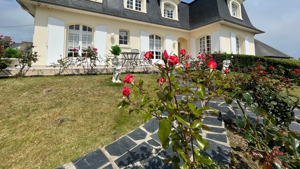 Una casa grande con fachada simétrica, múltiples ventanas y un jardín bien cuidado con rosales.