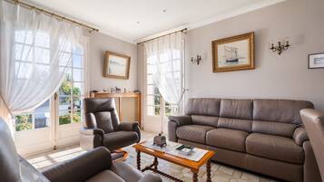 Ein Wohnzimmer mit braunem Ledersofa, einem braunen Ledersessel und einem Holztisch. Zwei Fenster mit durchsichtigen Vorhängen, ein gerahmtes Bild an der Wand und ein kleiner Tisch mit einem dekorativen Gegenstand.