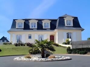 Une maison à deux étages, dotée d’une façade symétrique, d’une entrée centrale et d’un jardin aménagé à l’avant, avec un palmier et un élément décoratif circulaire en pierre.