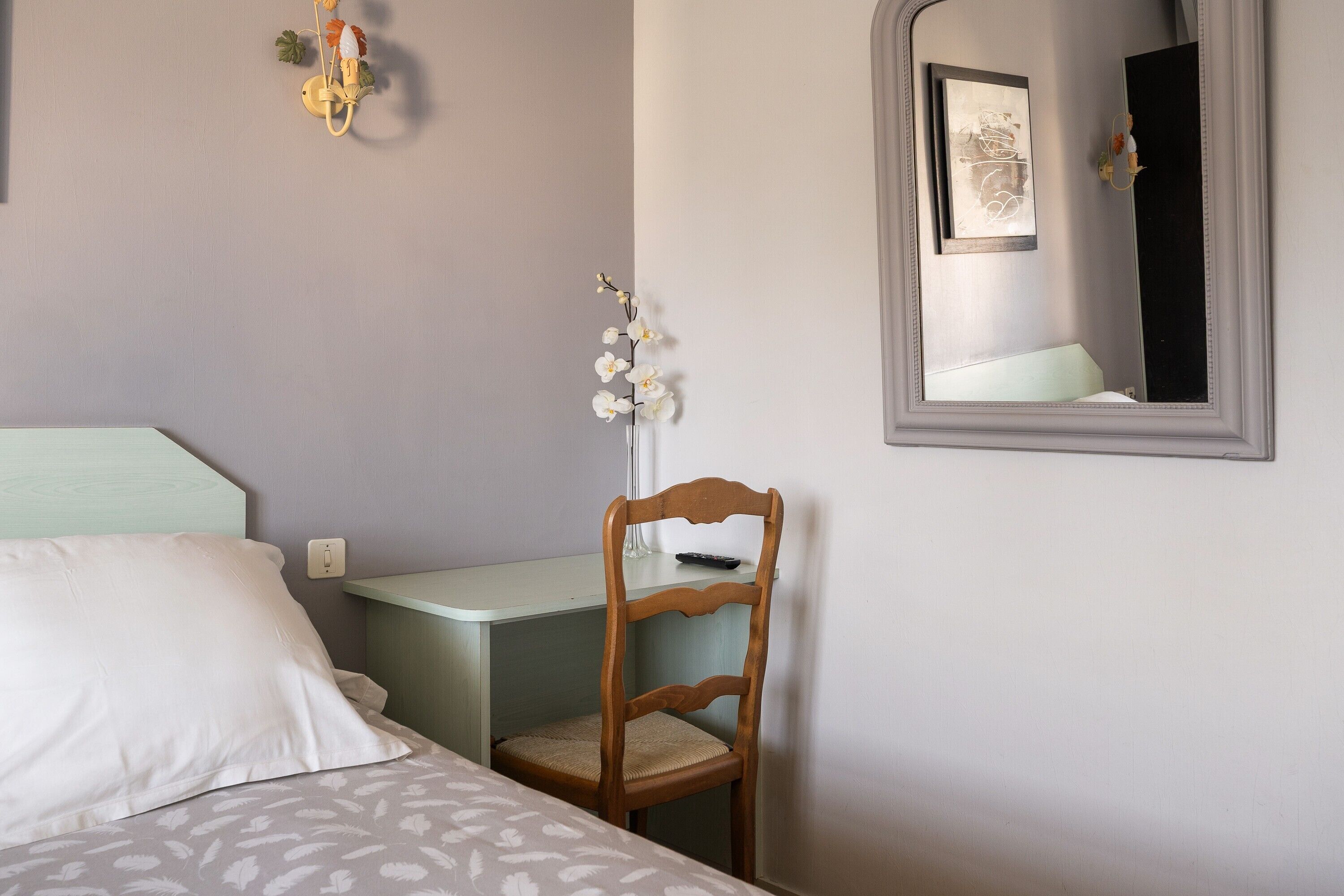 A bedroom with a bed, a desk, a chair, a mirror, and a wall-mounted light fixture.