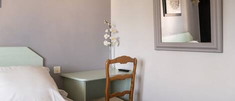 A bedroom with a bed, a desk, a chair, a mirror, and a wall-mounted light fixture.