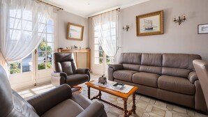Ein Wohnzimmer mit braunem Ledersofa, einem braunen Ledersessel und einem Holztisch. Zwei Fenster mit durchsichtigen Vorhängen, ein gerahmtes Bild an der Wand und ein kleiner Tisch mit einem dekorativen Gegenstand.