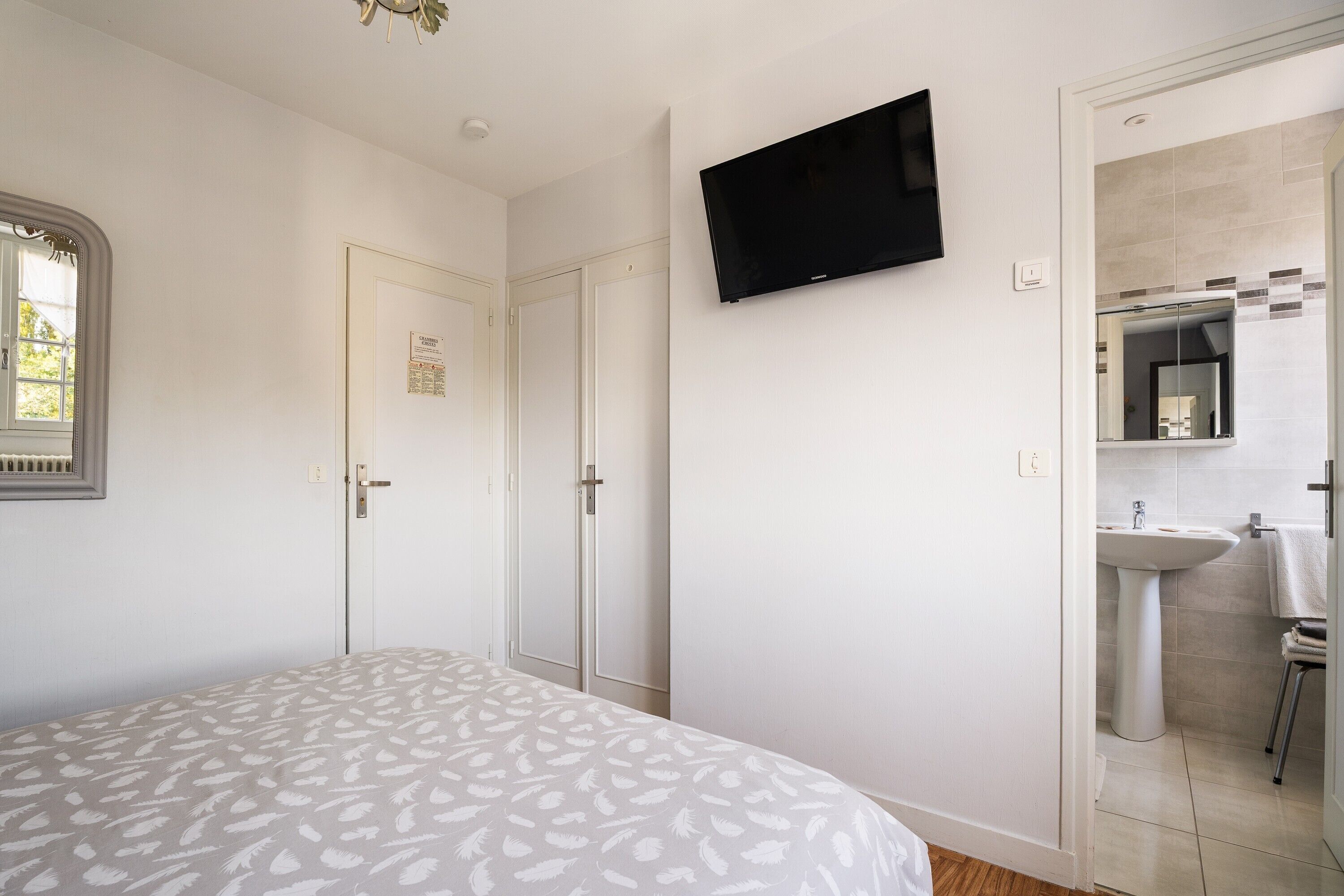 A bedroom with a bed, a TV mounted on the wall, and a bathroom visible through an open door.
