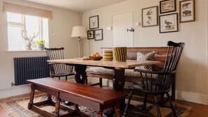 Un comedor con una mesa de madera, sillas negras, un banco, un radiador, una ventana con cortinas y obras de arte colgadas en la pared.