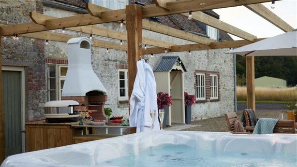 Un jacuzzi bajo una pérgola de madera, un edificio de piedra con chimenea y una pequeña cocina al aire libre.