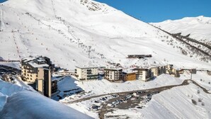 Un pueblo nevado en la montaña con varios edificios, un estacionamiento y pistas de esquí al fondo.