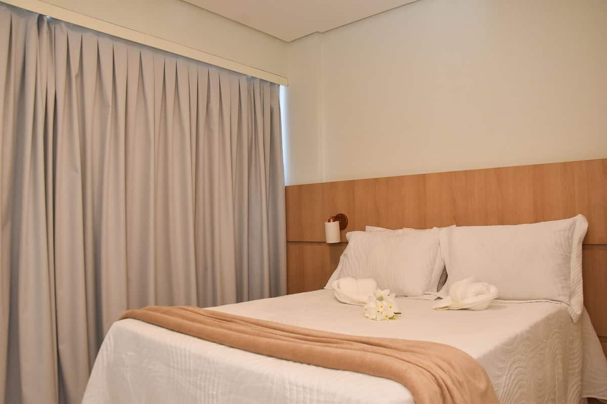 A hotel room with a bed, a wooden headboard, a bedside table with a lamp, and a window with curtains.