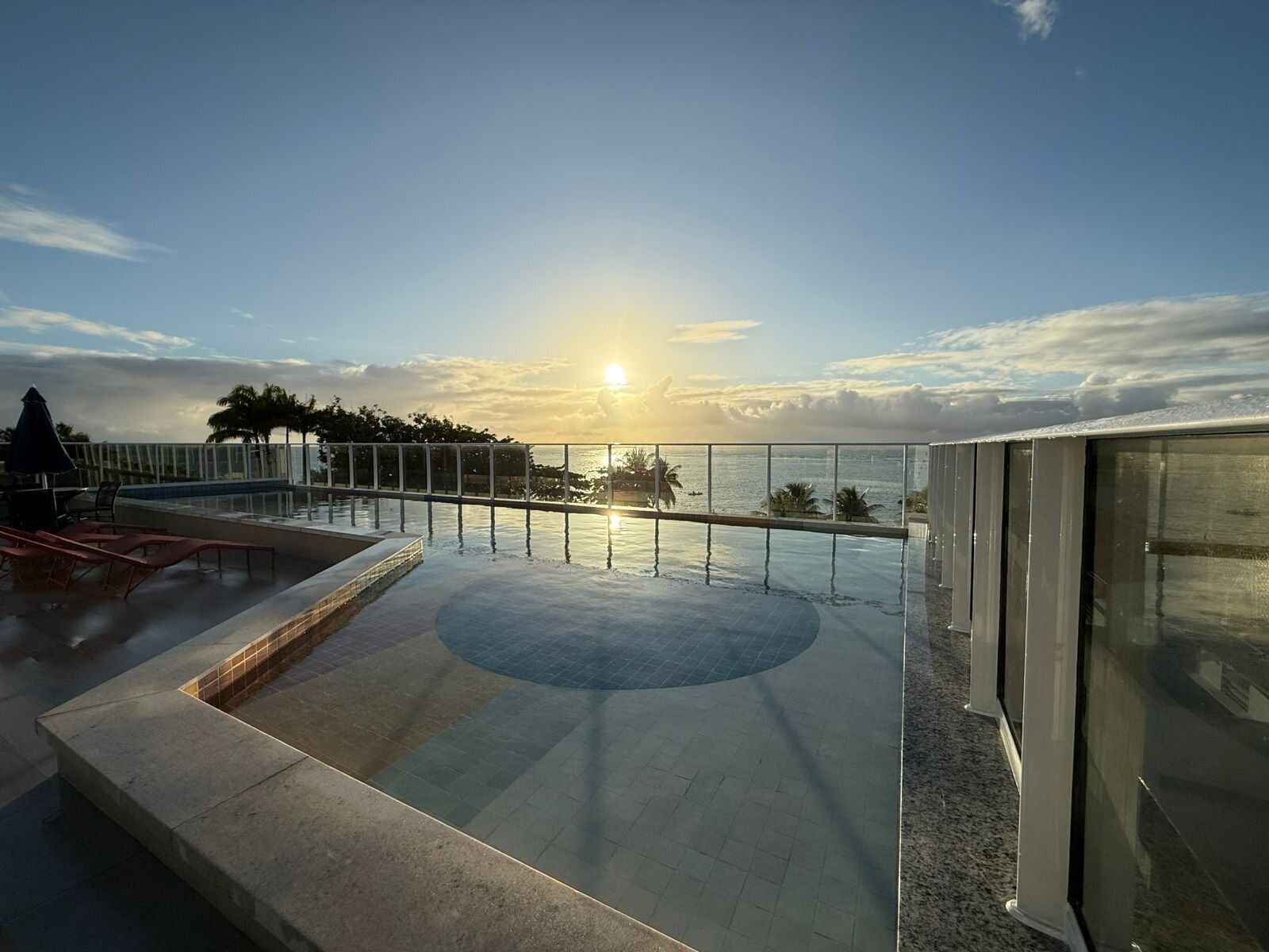 Una piscina a sfioro sul tetto con vista sul tramonto sull'oceano, una ringhiera in vetro e un'area relax con poltrone.