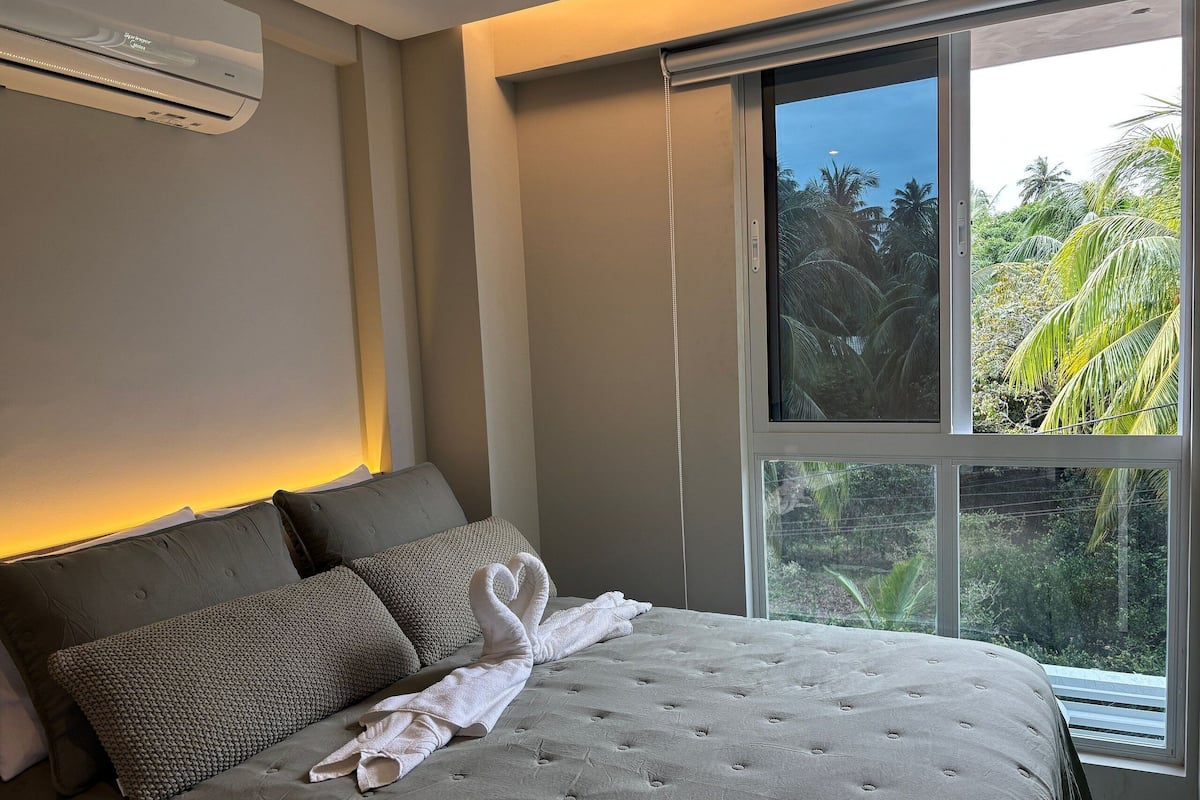A bedroom with a large window, a bed with a grey duvet, and a wall-mounted air conditioner.